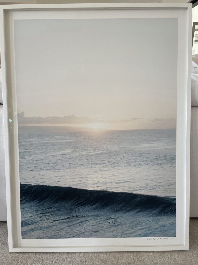 The artwork features a serene maritime scene with a tranquil wave gently rolling towards the viewer. The color palette is muted, dominated by cool tones that evoke a sense of calm and peace. Subtle gradients from the sky to the sea suggest an early morning or late afternoon ambiance, with a touch of warmth from the sun’s light gracing the horizon. The overall effect is both contemplative and soothing, inviting the observer to pause and reflect. The piece is in good condition, within a clean and simple white frame that complements its understated elegance. Hand signed Limited Edition Print number 6 out of 20, size is 30×40 inch titled “Honolua Wave” Kate Holstein is an American photographer who lives between Aspen, Colorado and the Caribbean island of Saint Barthélemy
