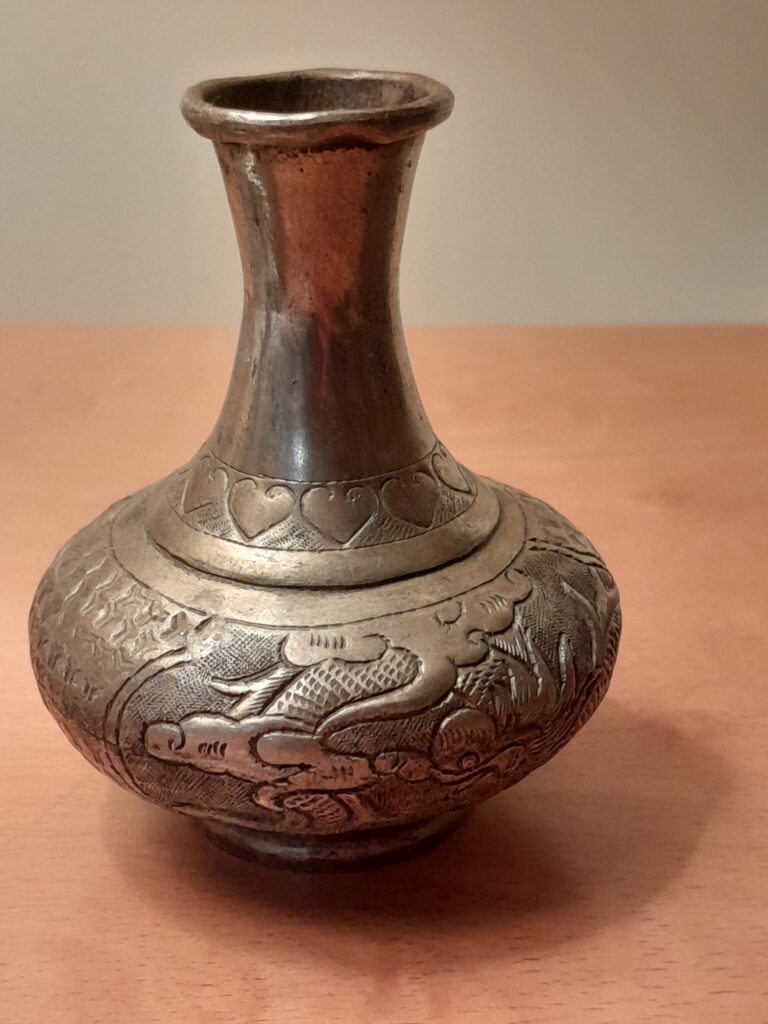 An antique metal vase (brass) with black patina (no copper) that suggests circa early 20thC. It features intricate engravings, possibly of an Asian-style dragon among clouds, which could indicate an origin in Chinese or similar East Asian cultures. The neck is slender, expanding to a bulbous body, typical of certain historical styles. It is a hand engraved vase with hand-joint pieces. Unmarked, origin South China.